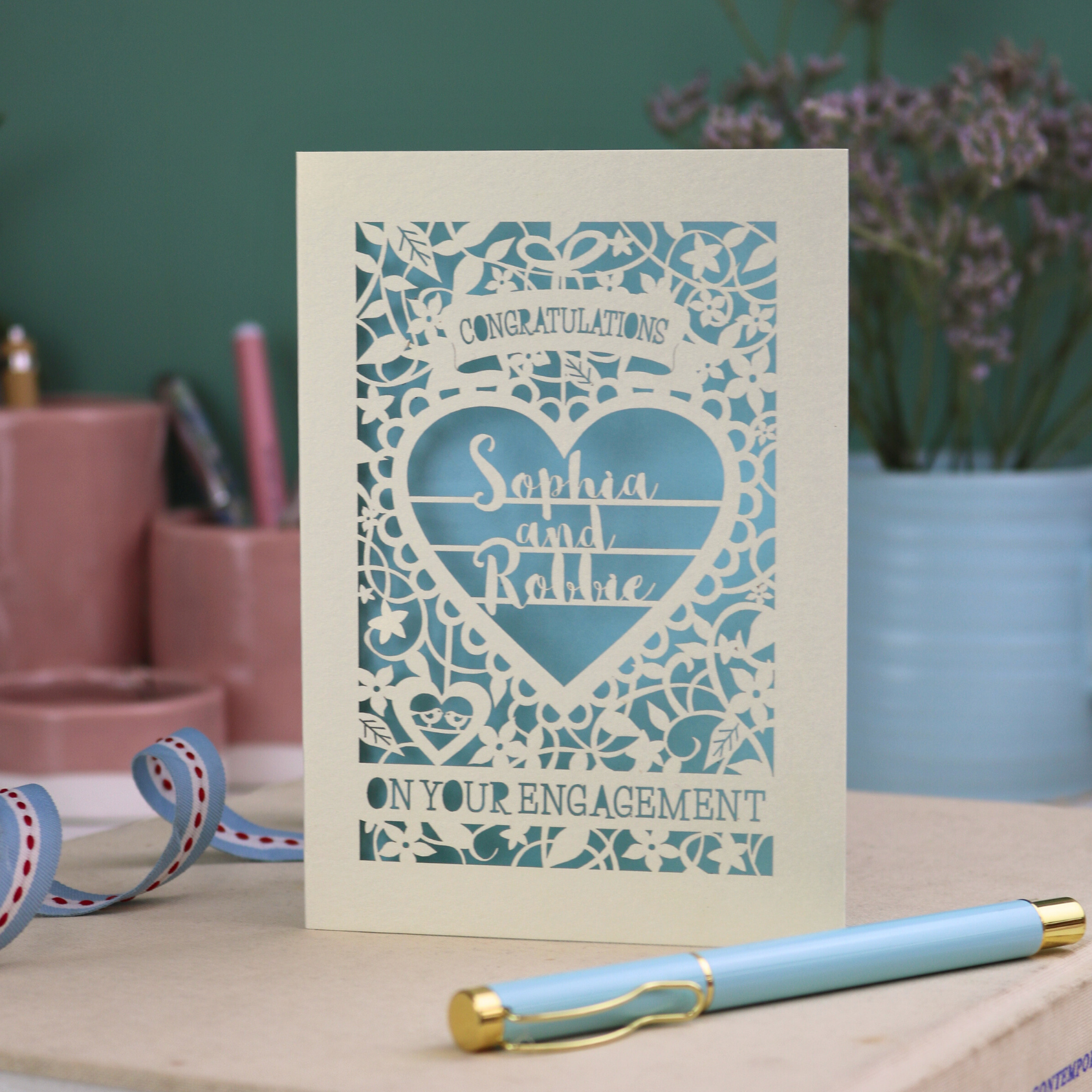 A cream laser cut engagement card on a desk with stationery items. The card is portrait-orientation, with floral cut out details and 'Congratulations, name and name, on your engagement'