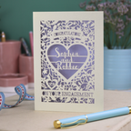 A cream laser cut engagement card on a desk with stationery items. The card is portrait-orientation, with floral cut out details and 'Congratulations, name and name, on your engagement'