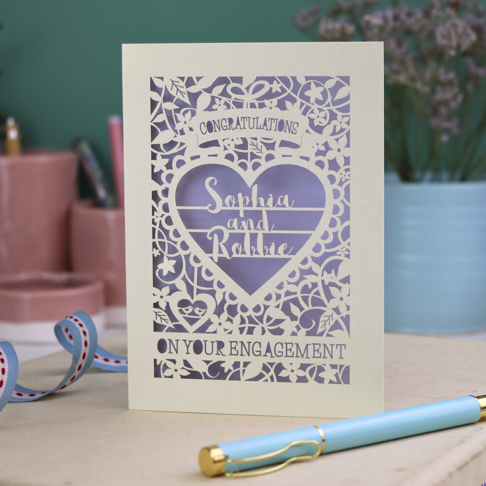 A cream laser cut engagement card on a desk with stationery items. The card is portrait-orientation, with floral cut out details and 'Congratulations, name and name, on your engagement'