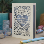 A cream laser cut engagement card on a desk with stationery items. The card is portrait-orientation, with floral cut out details and 'Congratulations, name and name, on your engagement'
