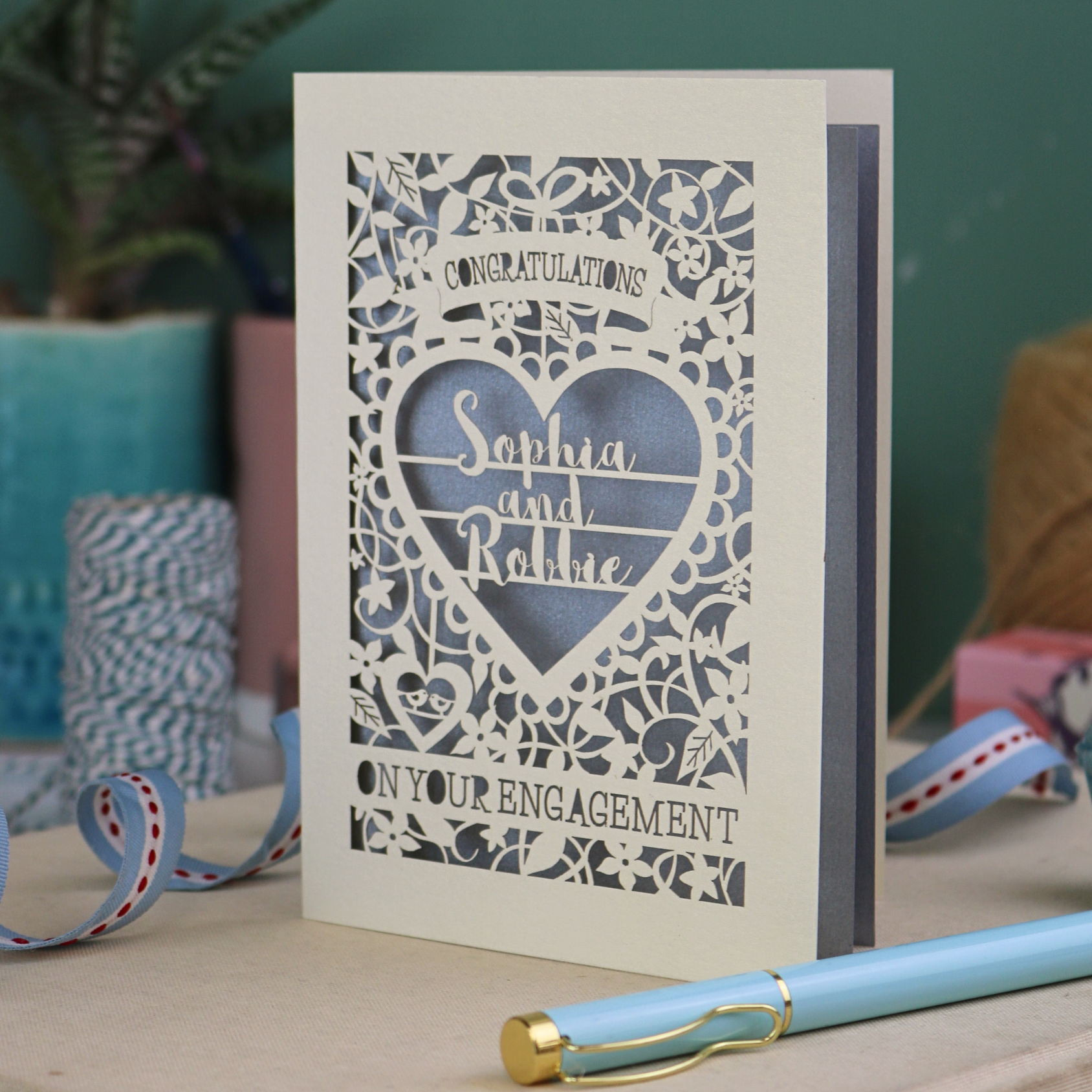 A cream laser cut engagement card on a desk with stationery items. The card is portrait-orientation, with floral cut out details and 'Congratulations, name and name, on your engagement'