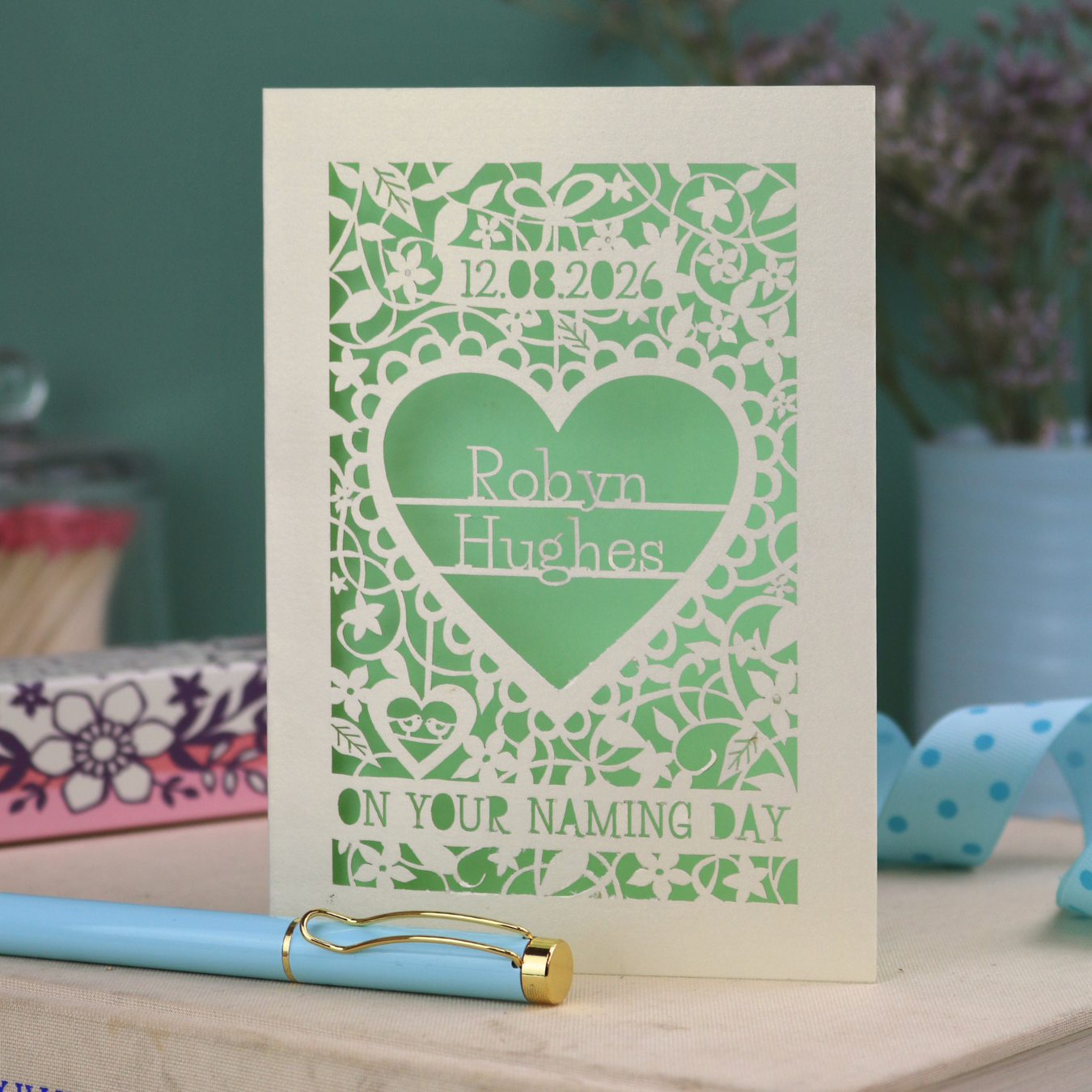 Personalised Naming Day card with decorative laser cut heart design and text on a wooden surface with a pen.