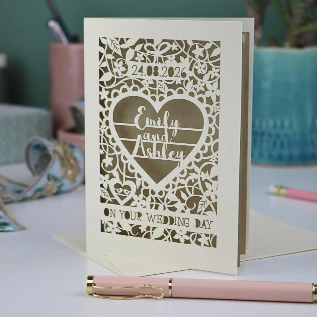 Personalised cut out wedding card with decorative patterns and names on a desk with stationery items.