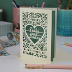 Personalised wedding card with decorative patterns and names on a desk with stationery items.