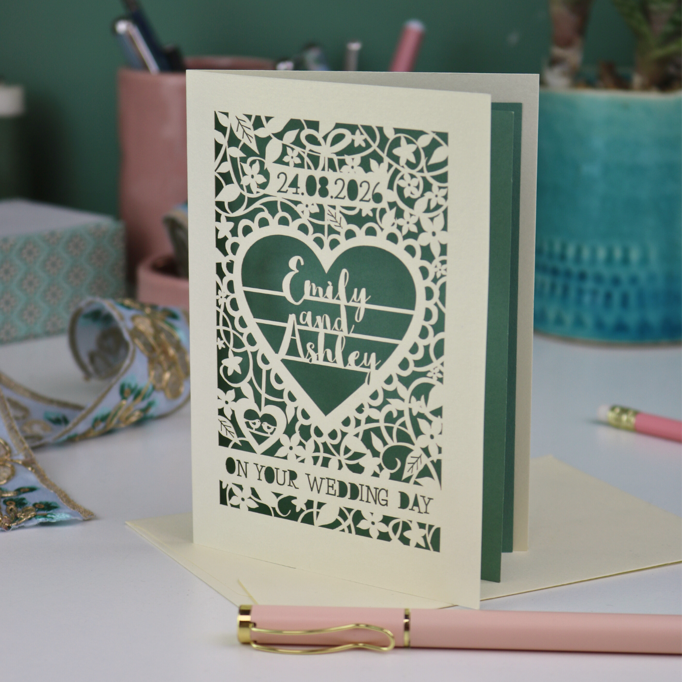 Personalised wedding card with decorative patterns and names on a desk with stationery items.