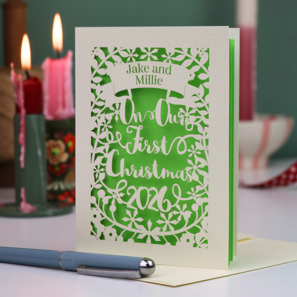 A personalized laser-cut Christmas card for partners, with cut out details over a bright green background, reading 'Jake and Millie Our First Christmas 2026'. The card is standing upright on a table with a blue pen in the foreground and blurred candles in the background.