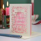 A personalized laser-cut Christmas greeting card with a pink and cream design, standing upright on a table with candles and a pen in the background.