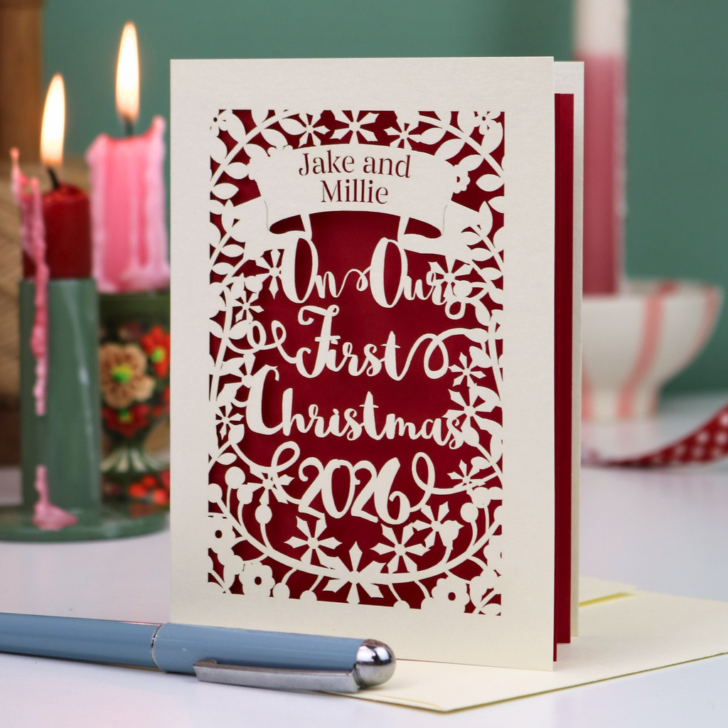 A personalized laser-cut Christmas greeting card with intricate cut out patterns, featuring the text 'Jake and Millie Our First Christmas 2026'. The card is styled on a tabletop with a blue pen in the foreground and lit candles in the background.