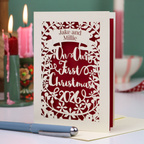 A personalized laser-cut Christmas greeting card with intricate cut out patterns, featuring the text 'Jake and Millie Our First Christmas 2026'. The card is styled on a tabletop with a blue pen in the foreground and lit candles in the background.