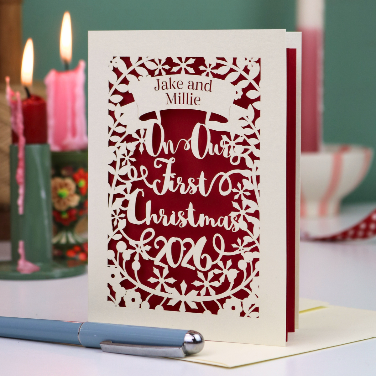 A personalized laser-cut Christmas greeting card with intricate cut out patterns, featuring the text 'Jake and Millie Our First Christmas 2026'. The card is styled on a tabletop with a blue pen in the foreground and lit candles in the background.