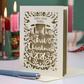 A personalized laser-cut Christmas card with intricate floral cut outs and the text 'Jake and Millie On Our First Christmas 2026', displayed on a table with candles in the background.