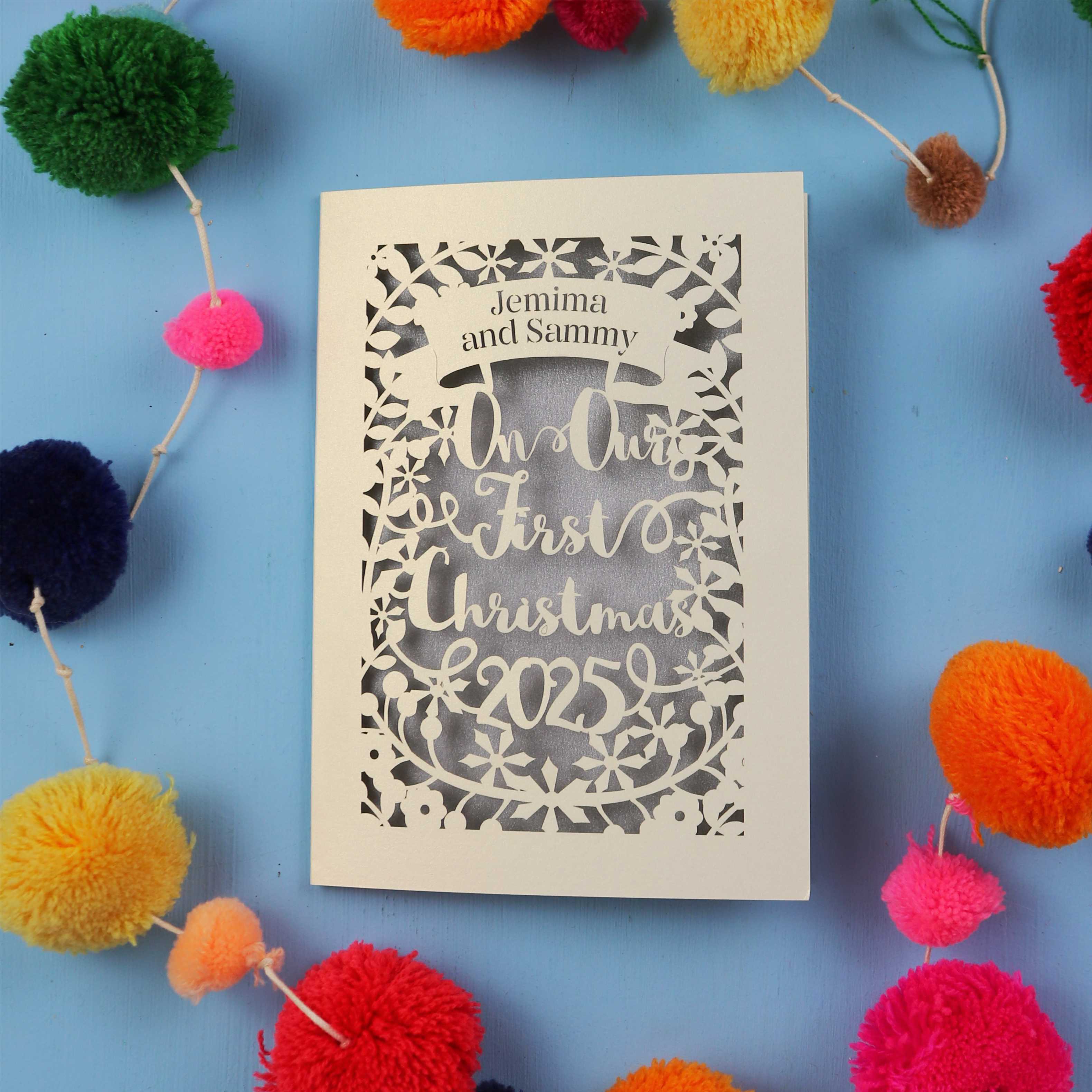 Personalized Christmas card with 'In Our First Christmas 2025' text, surrounded by colourful pom-poms on a blue background.