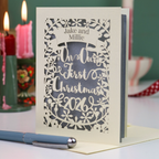 A personalized laser-cut Christmas card for a first Christmas together, standing upright on a table, with a blue pen in the foreground and blurred candles in the background. The card reads 'Jake and Millie On Our First Christmas 2026'.