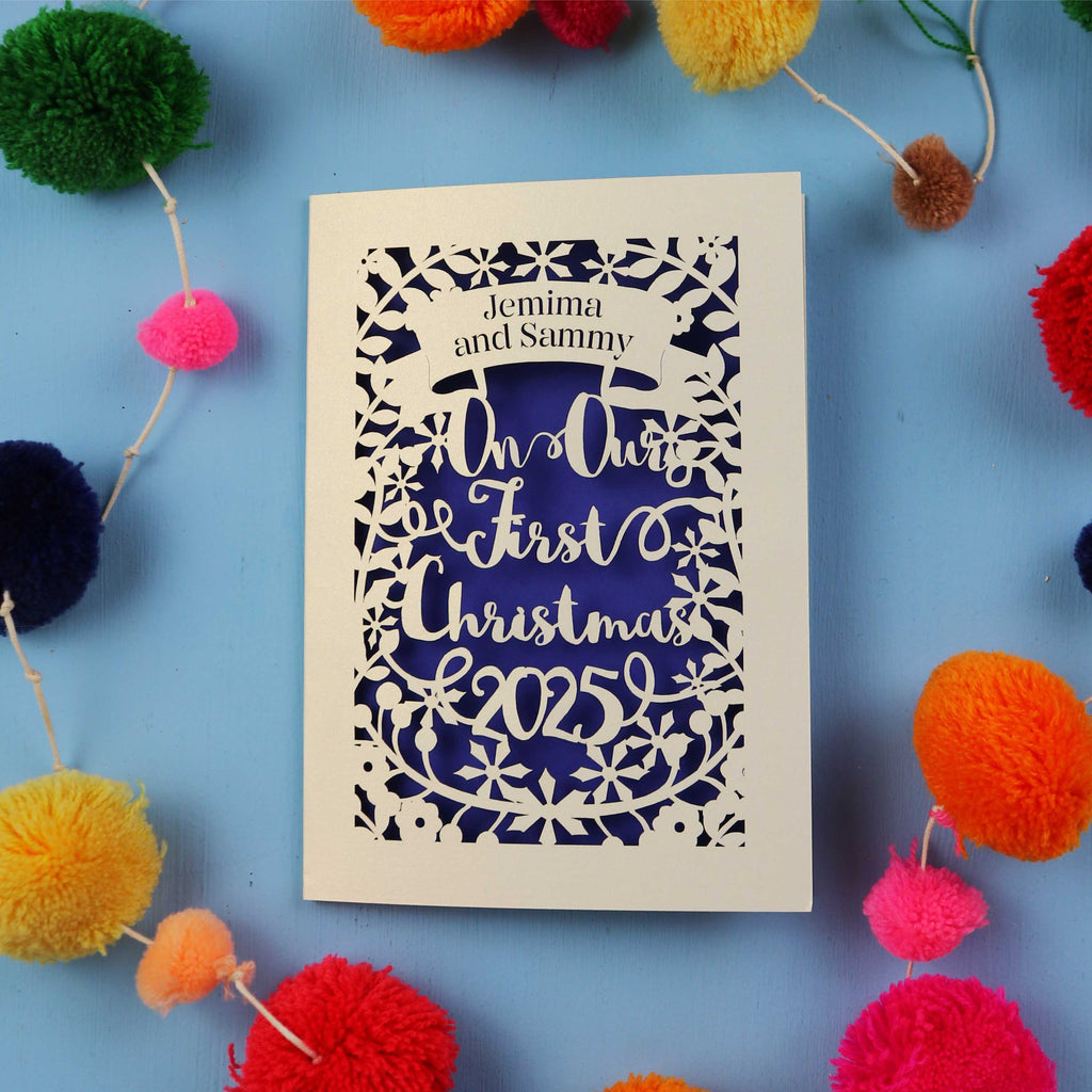 Personalized Christmas card with 'On Our First Christmas 2025' text, surrounded by colourful pom-poms on a blue background.