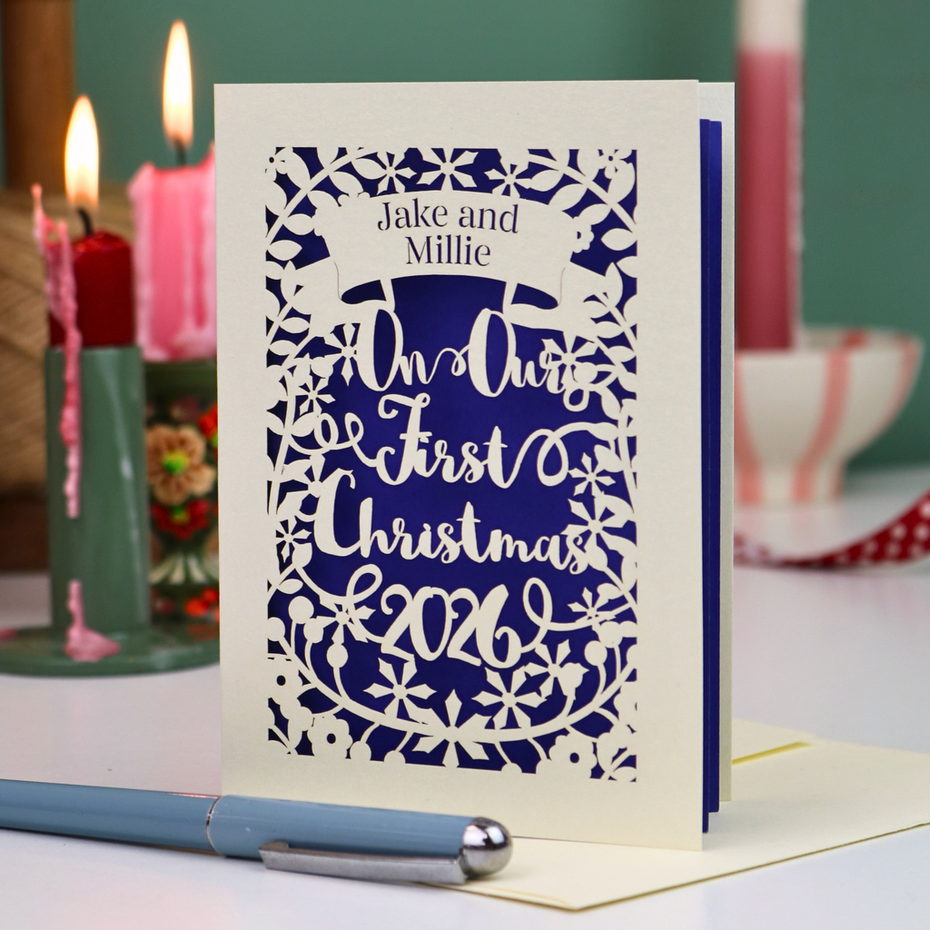 A personalized laser-cut Christmas greeting card with a cream border and violet paper backing, featuring the text 'Jake and Millie' and 'Our First Christmas 2026'. The card is displayed on a table with a blue pen in the foreground and lit candles in the background.