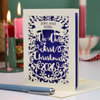 A personalized laser-cut Christmas greeting card with a cream border and violet paper backing, featuring the text 'Jake and Millie' and 'Our First Christmas 2026'. The card is displayed on a table with a blue pen in the foreground and lit candles in the background.