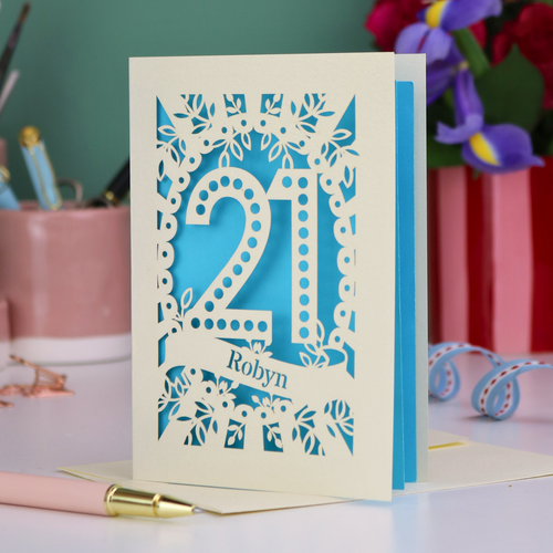 Special 21st birthday card with floral cut-out design and '21', personalised with a name in cut out stencil lettering, on a table with stationery items.