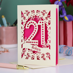 Unique 21st birthday card with floral cut-out design and '21', personalised with a name in cut out stencil lettering, on a table with stationery items.