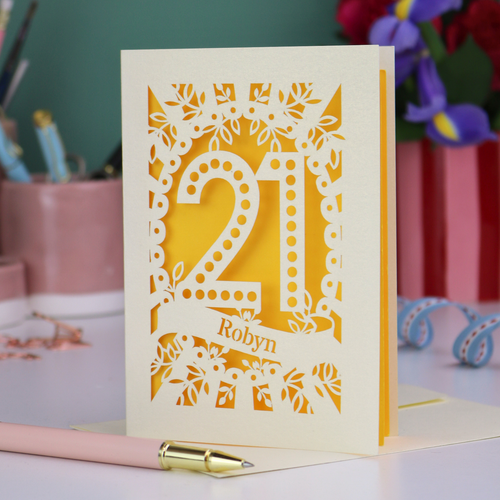 Decorative 21st birthday card with floral cut-out design and '21', personalised with a name in cut out stencil lettering, on a table with stationery items.