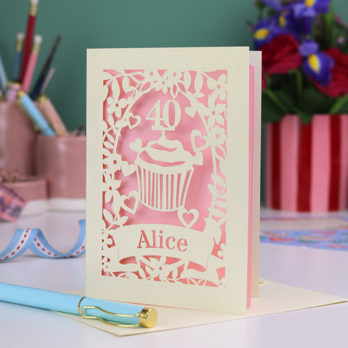 Decorative paper cut 40th birthday card with '40' and 'Alice', and a cupcake design, on a table with stationery items.