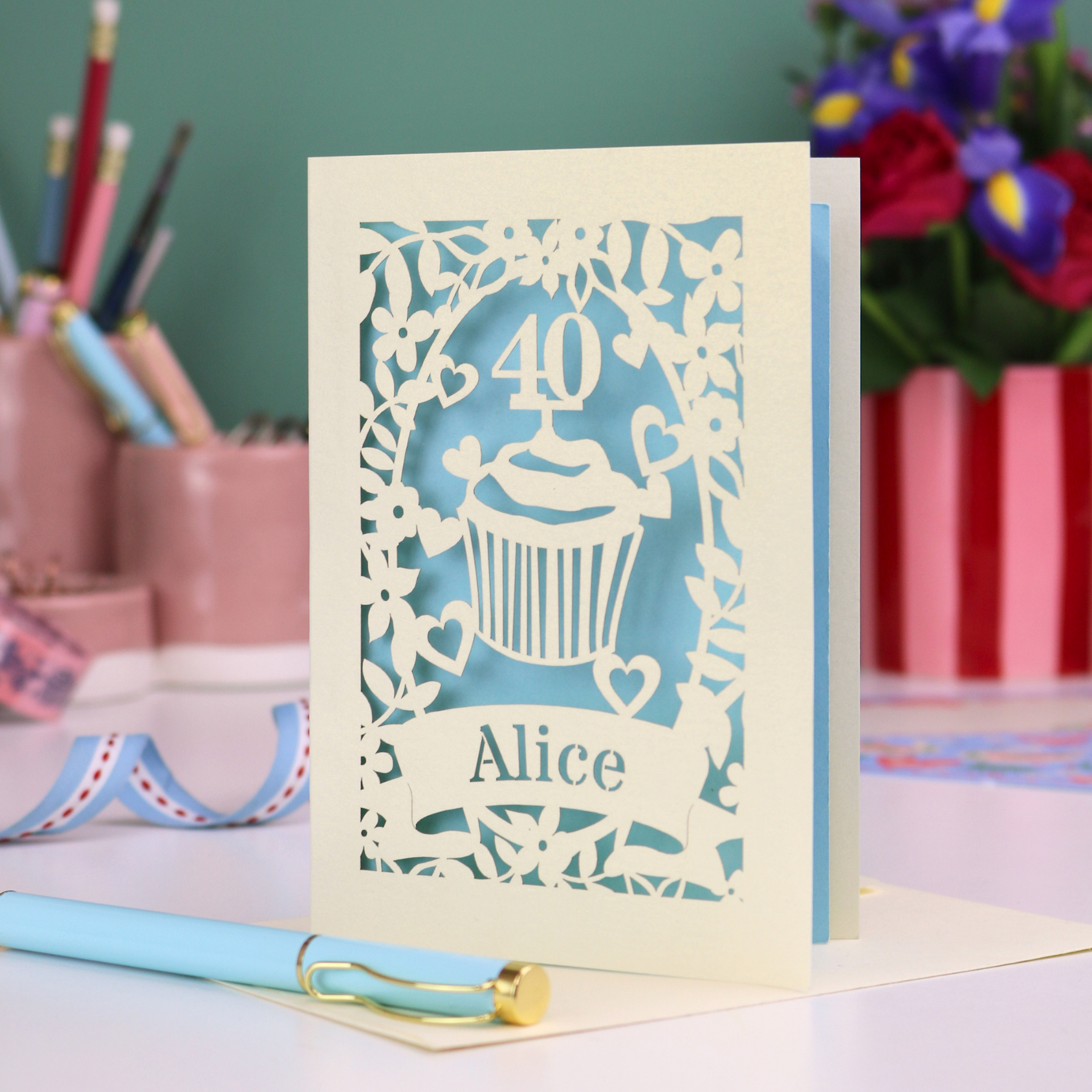 Decorative personalised 40th birthday card with '40' and 'Alice', and a cupcake design, on a table with stationery items.