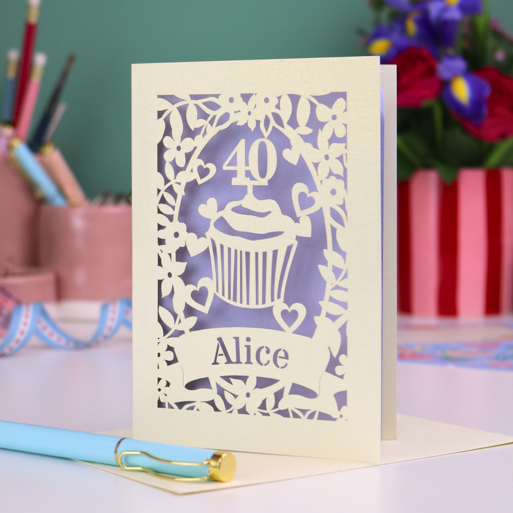 Decorative laser cut 40th birthday card with '40' and 'Alice', and a cupcake design, on a table with stationery items.