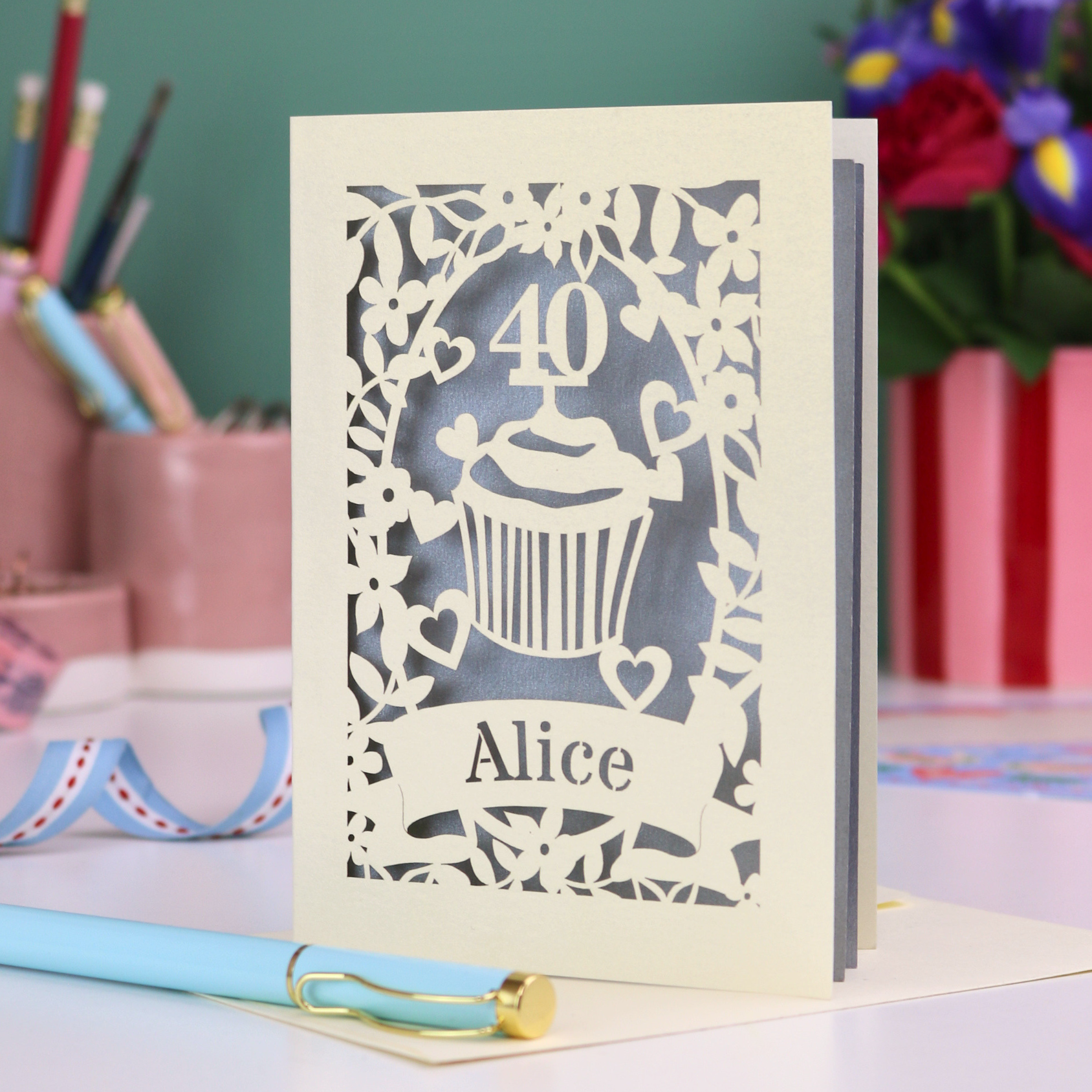 Decorative laser cut 40th birthday card with '40' and 'Alice', and a cupcake design, on a table with stationery items.