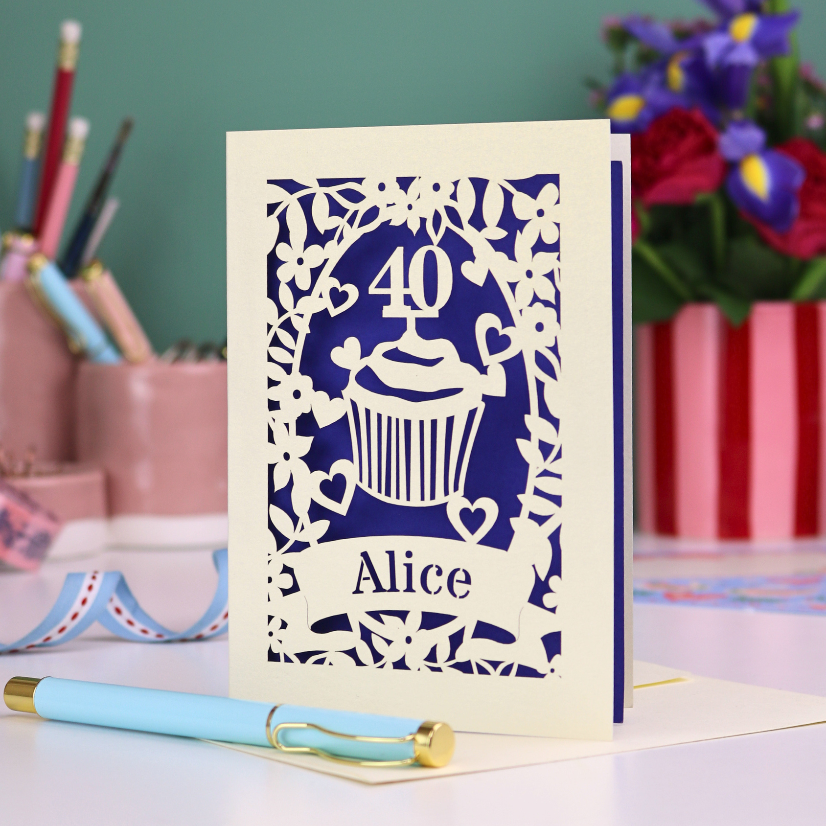 Decorative laser cut 40th birthday card with '40' and 'Alice', and a cupcake design, on a table with stationery items.