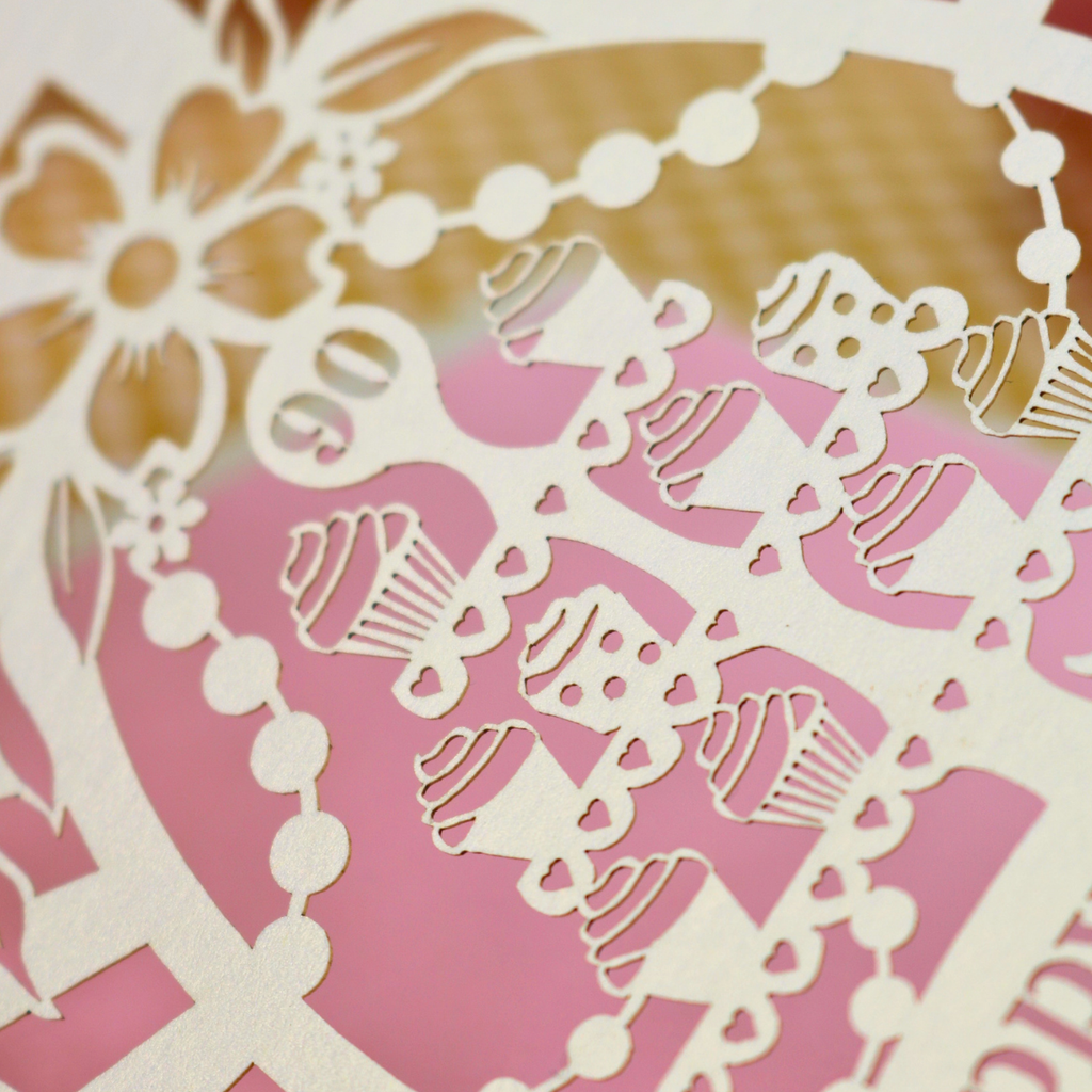 Close-up of a laser-cut decorative birthday card featuring cupcake and floral motifs over a pink and gold background.