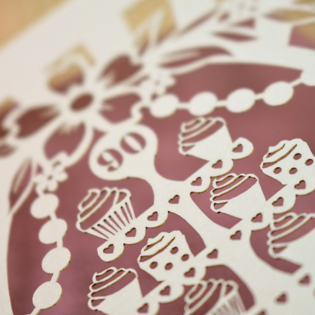 A close-up of a laser-cut paper decoration featuring cupcake and floral patterns over a dusty rose background.