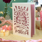 A personalized 90th birthday card with a laser-cut cupcake design and the text 'Happy Birthday Jenny' standing on a yellow-and-white checked tablecloth, surrounded by lit candles and pink ribbons.