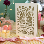 A personalized laser-cut birthday card with a tiered cake design and the text 'Happy Birthday Jenny' and '90', displayed on a yellow-and-white checked tablecloth with pink ribbons, lit candles, and a blurred festive background.