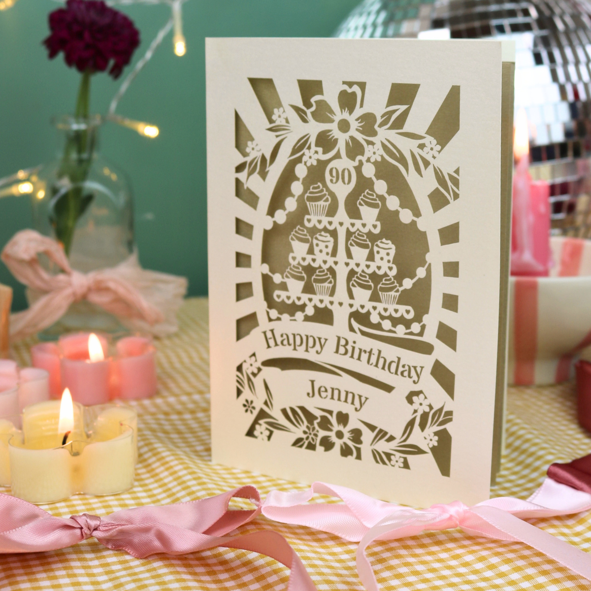 A personalized laser-cut birthday card with a tiered cake design and the text 'Happy Birthday Jenny' and '90', displayed on a yellow-and-white checked tablecloth with pink ribbons, lit candles, and a blurred festive background.