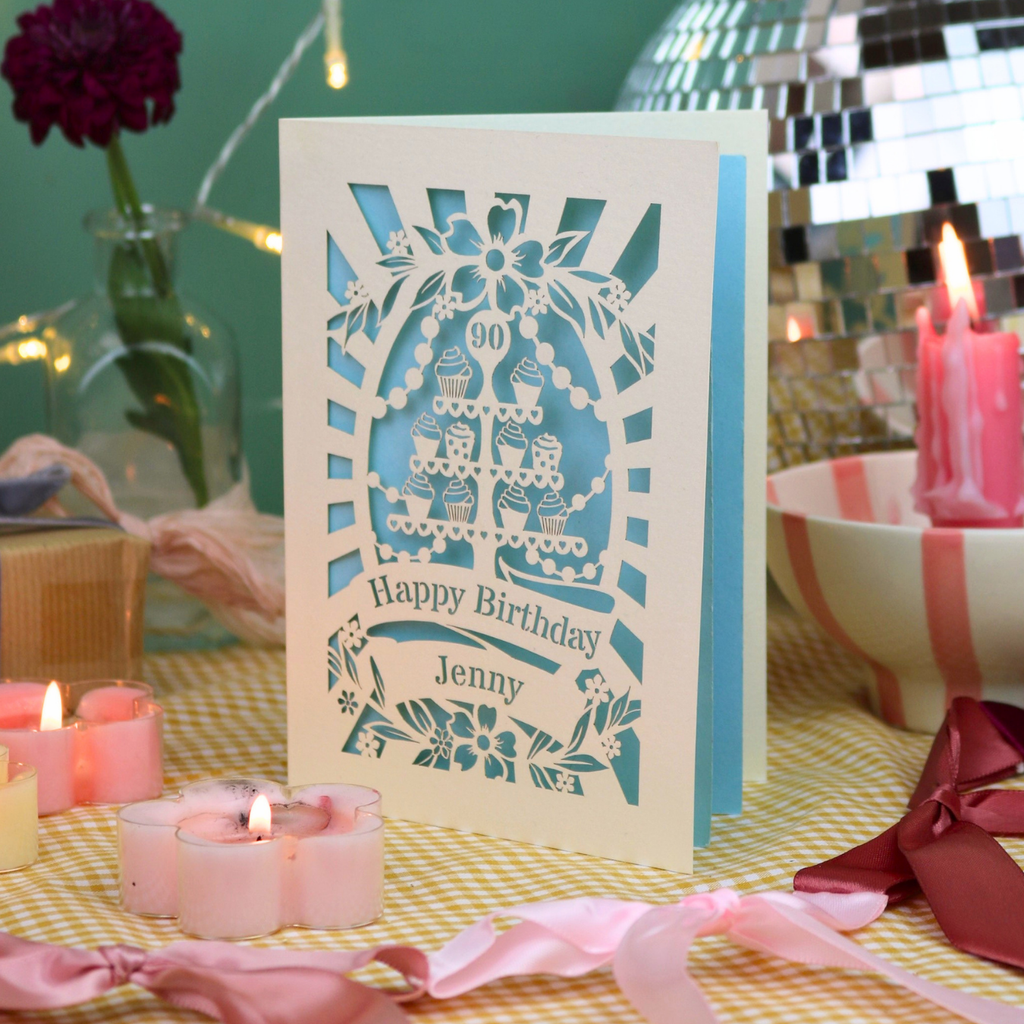 A personalized birthday card with a laser-cut white design showing cupcakes and the text 'Happy Birthday Jenny' and '90', displayed on a table with pink candles, ribbons, and a disco ball in the background.