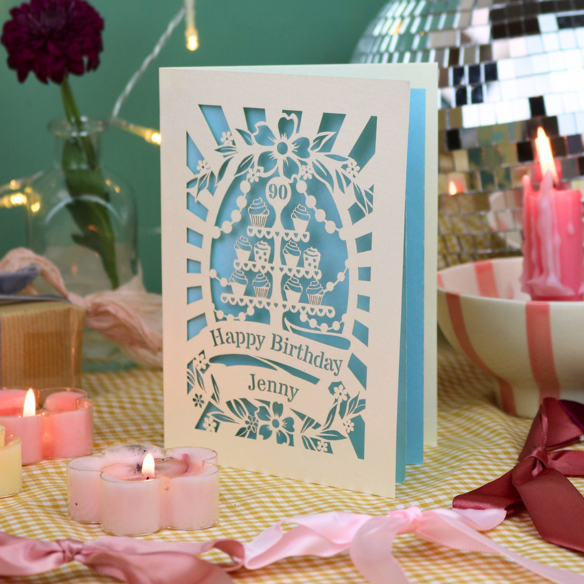 A personalized birthday card with a laser-cut white design showing cupcakes and the text 'Happy Birthday Jenny' and '90', displayed on a table with pink candles, ribbons, and a disco ball in the background.
