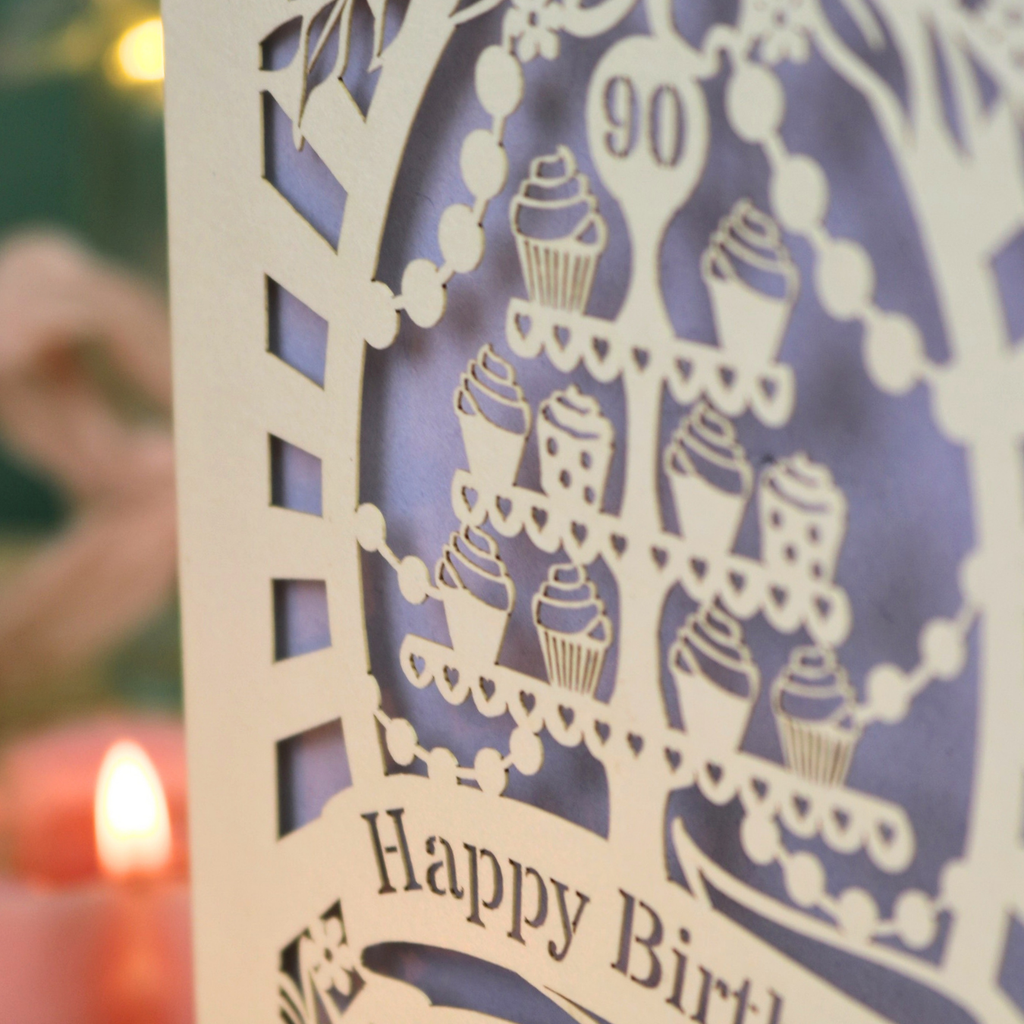A close-up of a laser-cut birthday card featuring a tiered cupcake stand, the number 90, and 'Happy Birthday' text, with a softly blurred candle and greenery in the background.