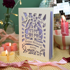 A personalised laser-cut birthday card with a cupcake design and the text 'Happy Birthday Jenny' and '90', displayed on a yellow-and-white checked tablecloth surrounded by lit pink and yellow candles, ribbons, and party décor.