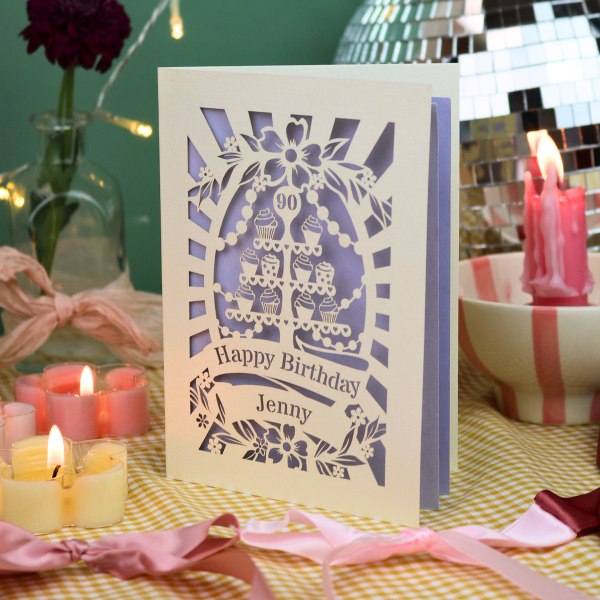 A personalised laser-cut birthday card with a cupcake design and the text 'Happy Birthday Jenny' and '90', displayed on a yellow-and-white checked tablecloth surrounded by lit pink and yellow candles, ribbons, and party décor.