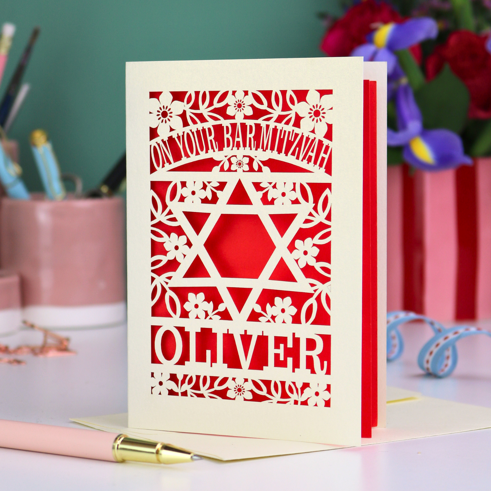 Personalized Bar Mitzvah card with cut out decorative design and name 'Oliver' on a desk with stationery items.