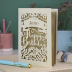 Personalised Laser cut birthday card with floral design and decorative "Happy Birthday" cut out text on a white surface with stationery items.