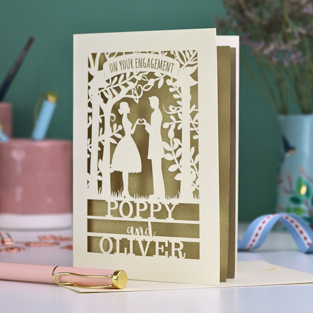 Engagement card with cut-out design of a couple, personalized with names 'Poppy' and 'Oliver', on a desk with stationery items.