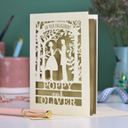 Engagement card with cut-out design of a couple, personalized with names 'Poppy' and 'Oliver', on a desk with stationery items.