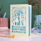 Laser cut Engagement card with cut-out design of a couple, personalized with 'Poppy and Oliver', on a desk with stationery items.