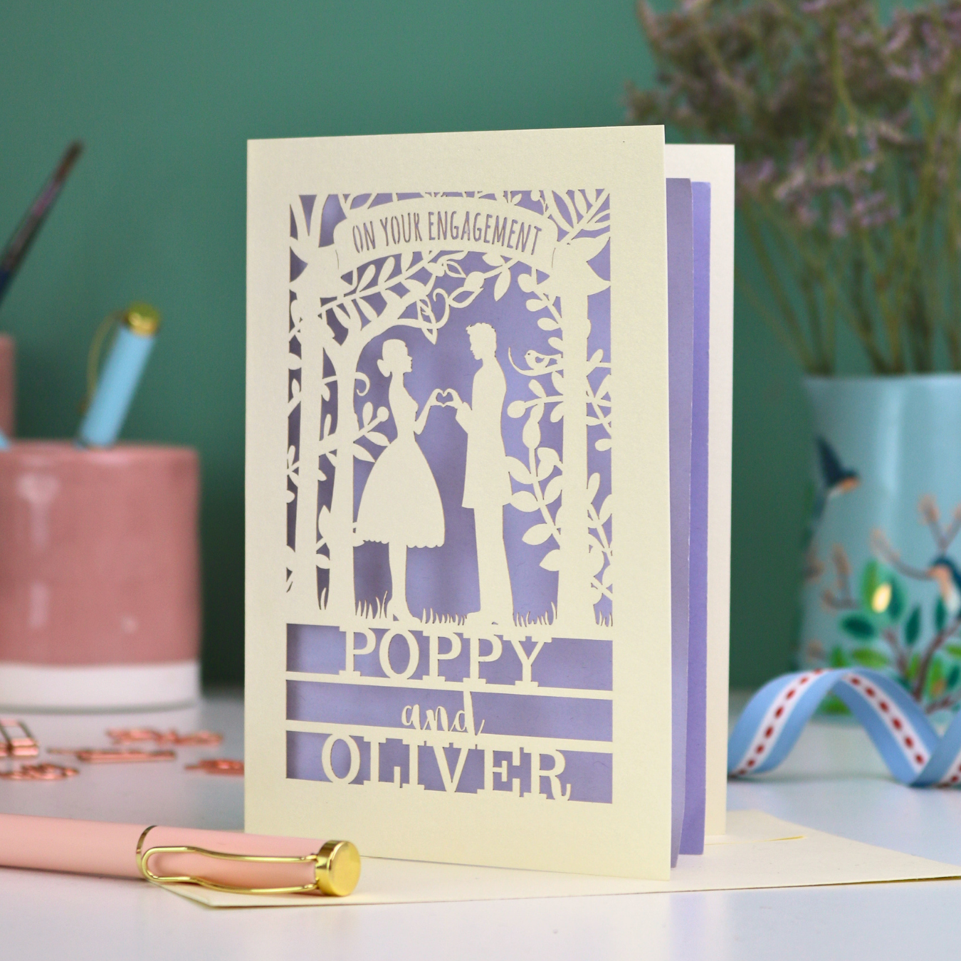 Engagement card with cut-out design of a couple, personalized with 'Poppy and Oliver', on a table with stationery items.