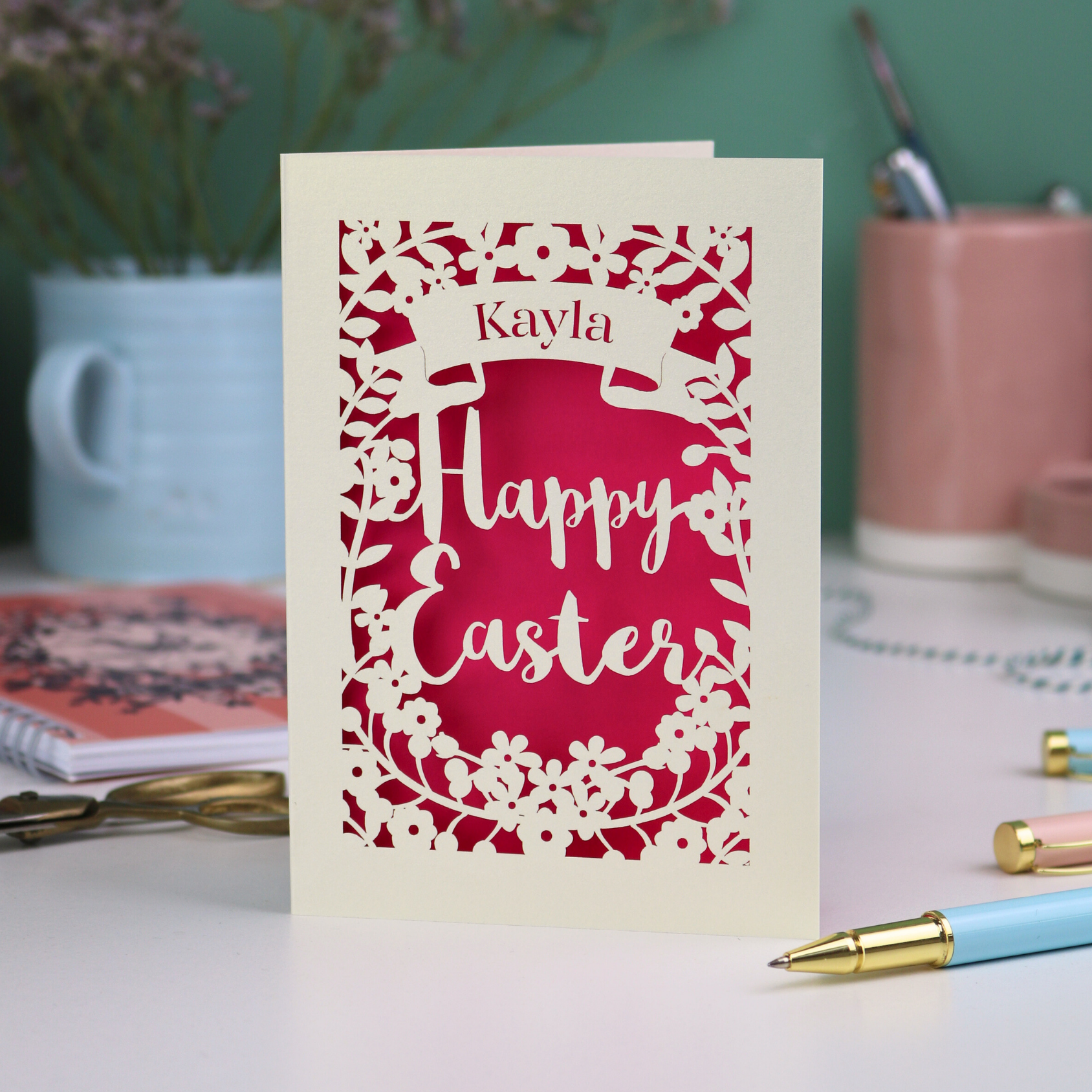 Easter card with 'Happy Easter' and 'Kayla' text on a pink background, placed on a desk with stationery items.