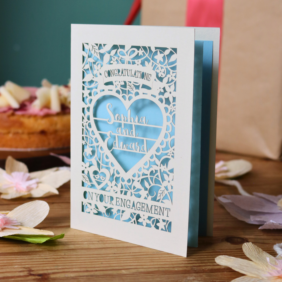 A cream laser cut engagement card with intricate floral border and heart design. Inside the heart are the names 'Sophia and Edward' in a brush script font, above is the word 'Congratulations', and below 'on your engagement'