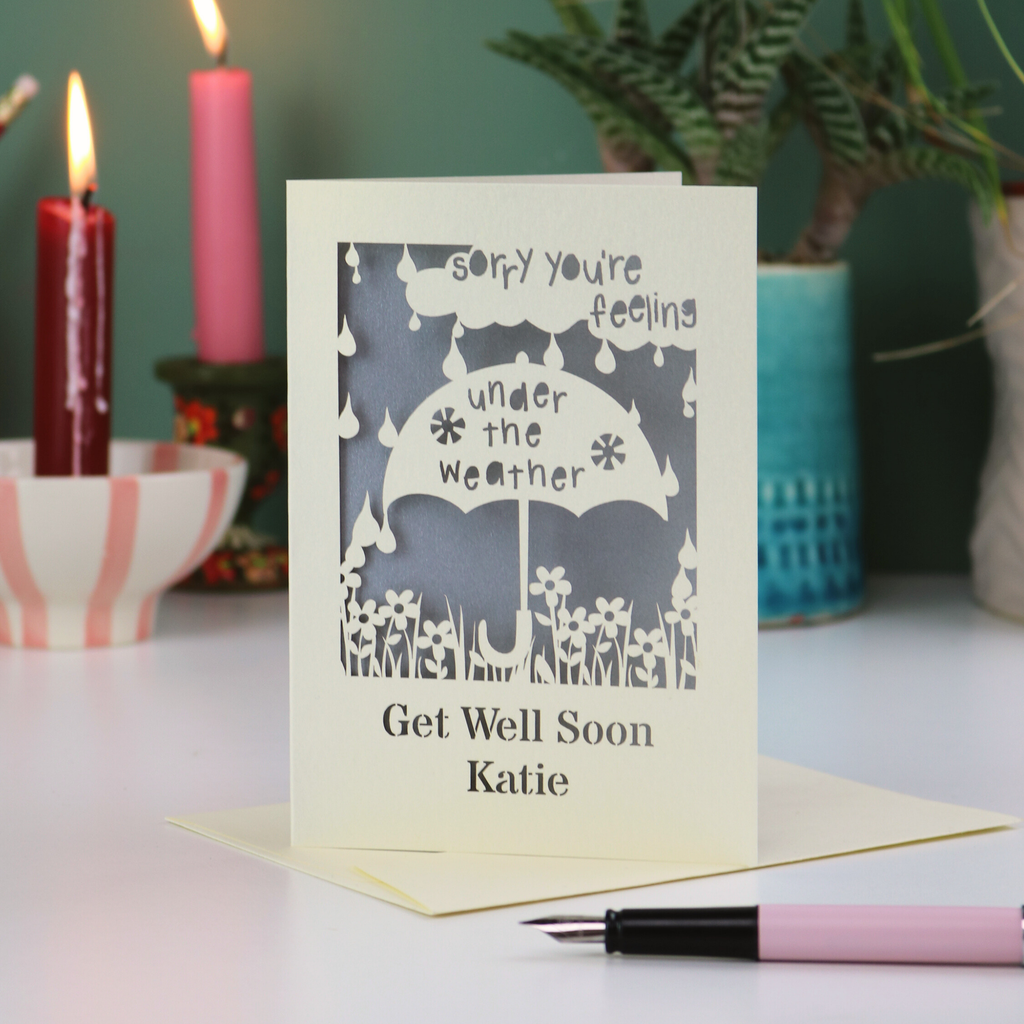 A laser cut get well soon card, with cut out umbrella design and the text 'Sorry you're feeling under the weather, get well soon' and personalised with a name.