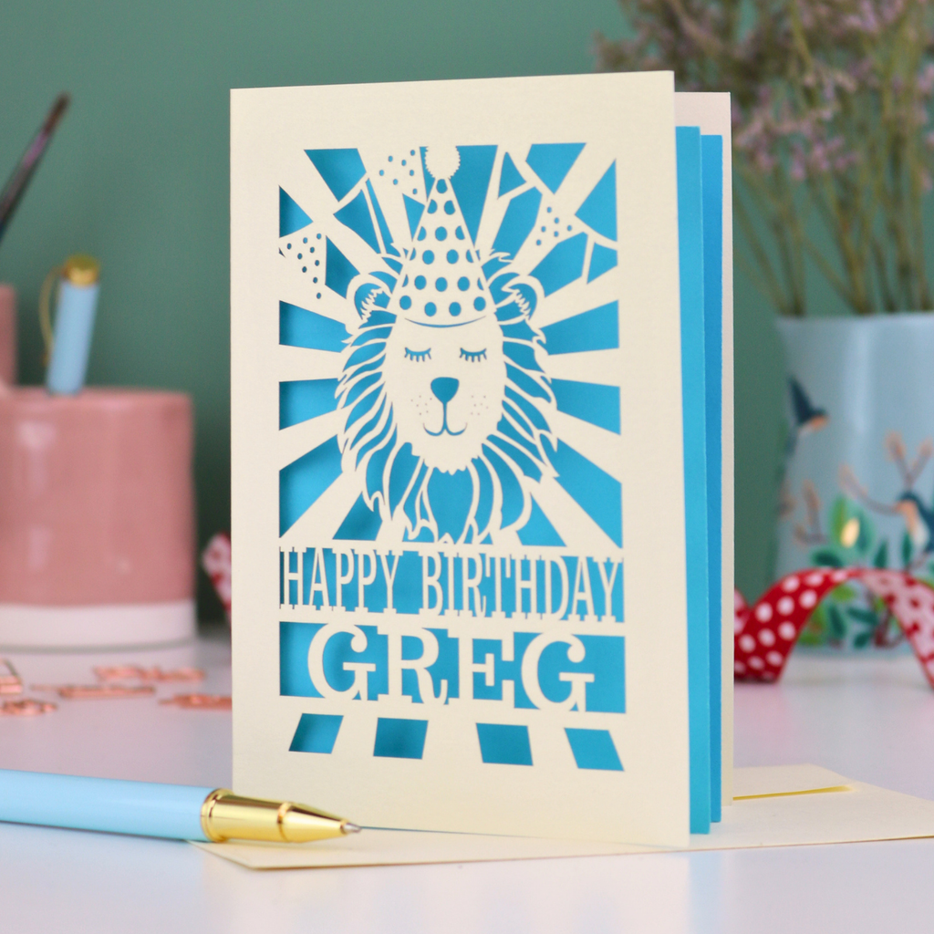 A personalised birthday card with laser cut lion in a party hat design and personalised text "Happy Birthday Greg", on a white desk with pen and stationery items