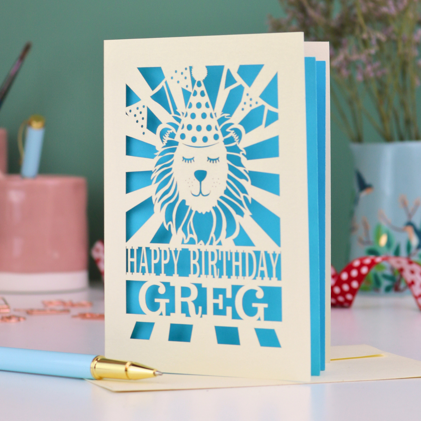 A personalised birthday card with laser cut lion in a party hat design and personalised text "Happy Birthday Greg", on a white desk with pen and stationery items