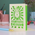 A personalised birthday card with laser cut lion in a party hat design and personalised text "Happy Birthday Greg", on a white desk with pen and stationery items